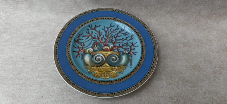 May include: A decorative plate with a blue border and a central design featuring coral, seashells, and a stylized shell-like structure. The plate has a gold and black patterned trim and a light blue center.