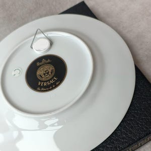 May include: White porcelain decorative plate with a black and gold Versace logo on the back. The plate has a metal hanger attached for display. The plate is resting on a black textured surface.