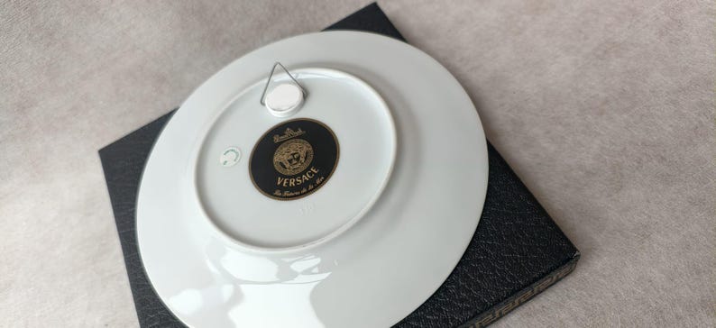 May include: White porcelain decorative plate with a black and gold Versace logo on the back. The plate has a metal hanger attached for display. The plate is resting on a black textured surface.