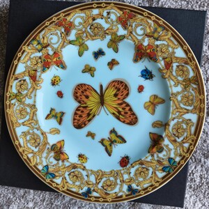 May include: A decorative plate with a light blue centre and a gold rim. The plate features a butterfly and floral design with butterflies in various colours, including orange, yellow, blue, and green. The plate is on a black surface.