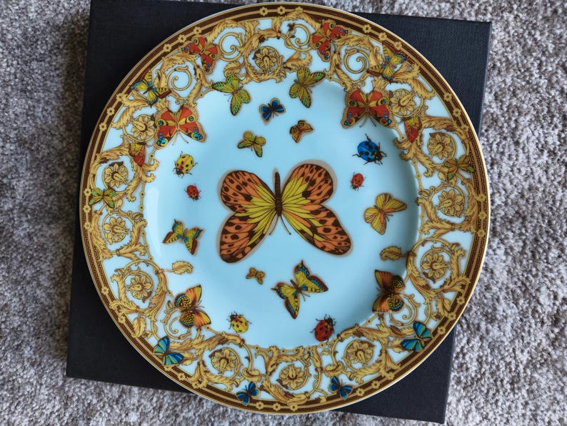 May include: A decorative plate with a light blue centre and a gold rim. The plate features a butterfly and floral design with butterflies in various colours, including orange, yellow, blue, and green. The plate is on a black surface.