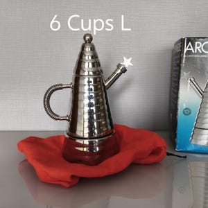 May include: A stainless steel Archimede espresso maker with a star-shaped spout, sitting on a red cloth. The box next to it reads "ARCHIMEDE". The text "6 Cups L" is above the espresso maker.