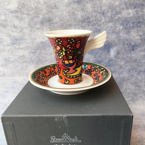 May include: A decorative coffee cup and saucer set with a vibrant floral and bird design. The cup has a wing-shaped handle and sits on a matching saucer. The set is presented on a black box with the "Rosenthal studio-linie" logo.
