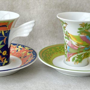 May include: Two decorative porcelain teacups with matching saucers. One cup features a blue floral design with a white wing-shaped handle. The other cup has a colorful bird and leaf design with a yellow wing-shaped handle. Both cups sit on white saucers.