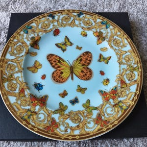 May include: A decorative plate with a light blue background, featuring a butterfly and insect design. The plate has a gold rim and ornate gold detailing around the edge. Butterflies in various colours are scattered across the surface.