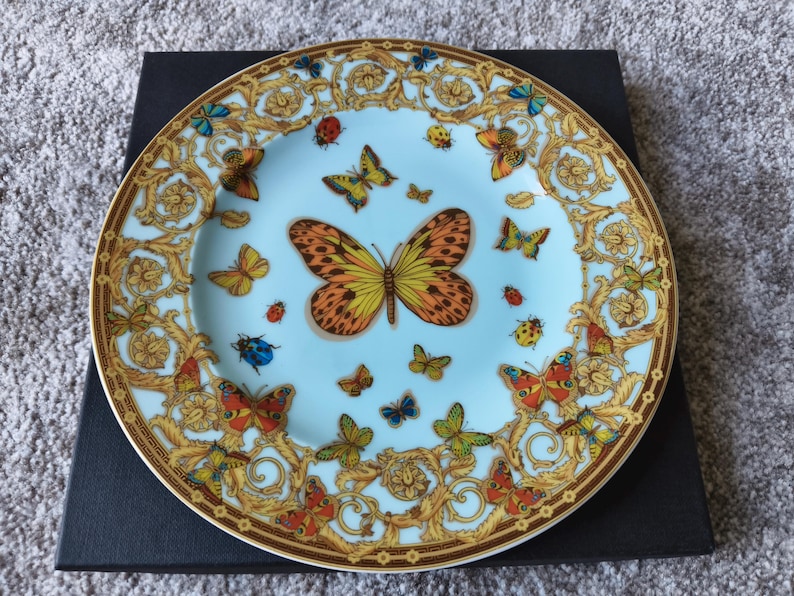 May include: A decorative plate with a light blue background, featuring a butterfly and insect design. The plate has a gold rim and ornate gold detailing around the edge. Butterflies in various colours are scattered across the surface.