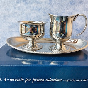 May include: A polished silver breakfast set comprising a small jug with a curved handle and a cup, presented on an oval tray. The set is made of shiny metal and rests on a dark blue surface. The text "art. 4 - servizio per prima colazione - acciaio inox 18/10" is visible.