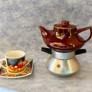 May include: A brown ceramic teapot sits atop a silver stovetop espresso maker. A decorative teacup and saucer with a fruit design are to the left. The items are set against a textured, light blue background.