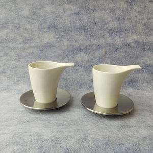 May include: Two matching cream-colored ceramic cups with a spout, resting on round, silver-colored saucers. The cups have a modern, minimalist design. The background is a light blue, textured surface.
