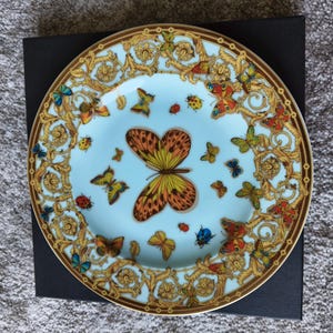 May include: A decorative plate with a light blue centre featuring a large butterfly and several smaller butterflies in various colours. The plate has a gold and brown ornate border with butterfly and insect designs.