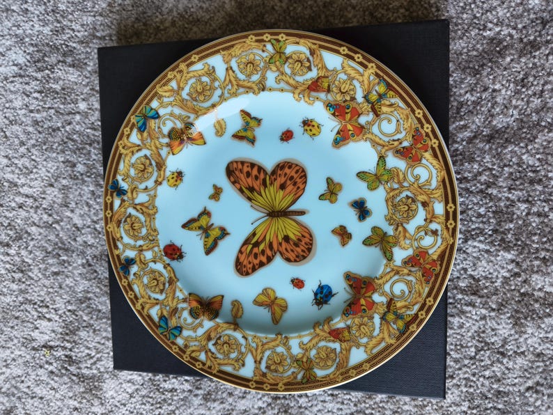 May include: A decorative plate with a light blue centre featuring a large butterfly and several smaller butterflies in various colours. The plate has a gold and brown ornate border with butterfly and insect designs.