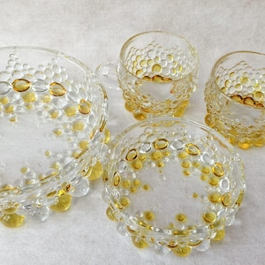 Könnte beinhalten: Ein klares Glasservice mit strukturiertem Blasendesign. Das Set umfasst eine große Schüssel, zwei kleine Schalen und zwei Tassen mit Henkeln. Die Blasen sind mit bernsteinfarbenem und klarem Glas akzentuiert.
