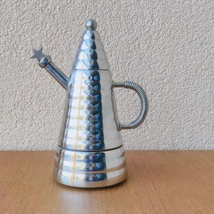 Op de afbeelding: Een zilverkleurige espressomaker met een gehamerde textuur. Het heeft een conische vorm, een kleine tuit met een stervormig detail en een lusvormige handgreep. De espressomaker staat op een houten oppervlak.