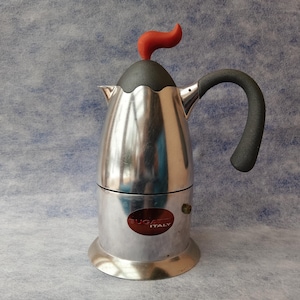 May include: A stainless steel stovetop espresso maker with a black handle and a red accent on top. The coffee maker has a round base and a label that reads "BUGA ITALY". The espresso maker is designed for making coffee.