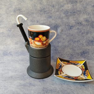 May include: A coffee cup with a fruit design sits on a dark grey stand with a white spout. A matching square saucer with a yellow, black, and blue pattern is next to the cup. The cup and saucer are on a light blue surface.
