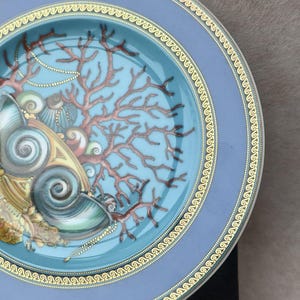 May include: A decorative plate with a blue rim and a central design featuring seashells, coral, and pearls. The plate has a gold border and is set against a neutral background. The plate's design is detailed and colorful.