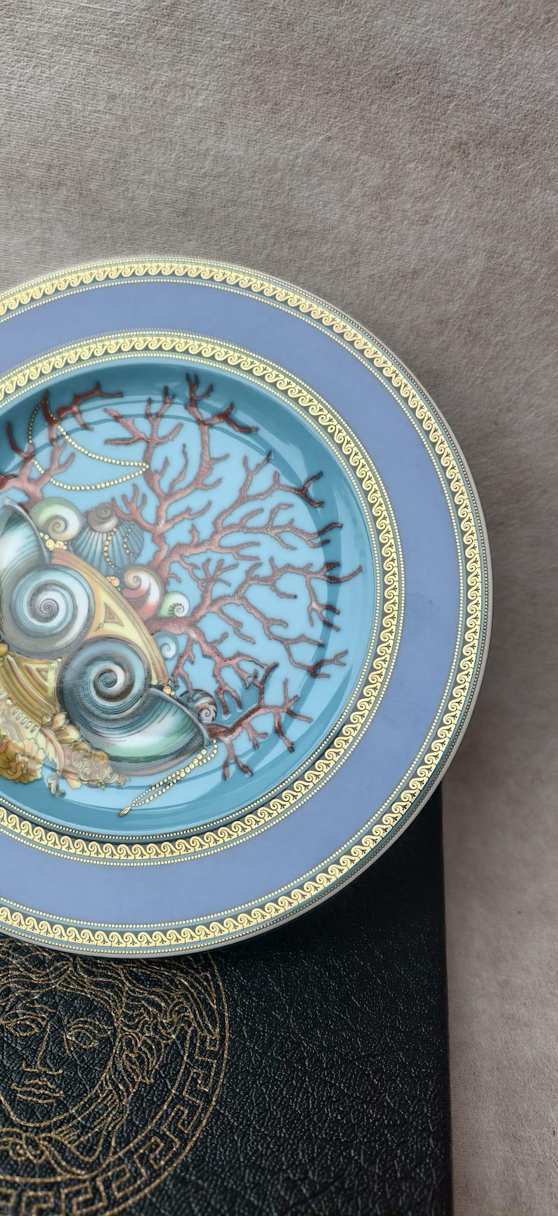 May include: A decorative plate with a blue rim and a central design featuring seashells, coral, and pearls. The plate has a gold border and is set against a neutral background. The plate's design is detailed and colorful.