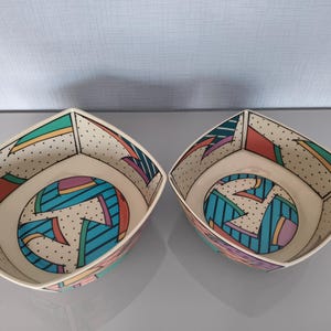 Könnte beinhalten: Zwei quadratische Keramikschalen mit cremefarbener Basis und schwarzem Punktmuster. Die Innenflächen sind mit geometrischen Formen in Türkis, Pink, Rot und Blau verziert, die durch kräftige schwarze Umrandungen und Linien betont werden.