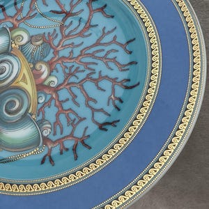 May include: Decorative ceramic plate with a blue and gold rim. The plate features a turquoise center with a coral and seashell design. The coral is brown, and the seashells are beige and gold.