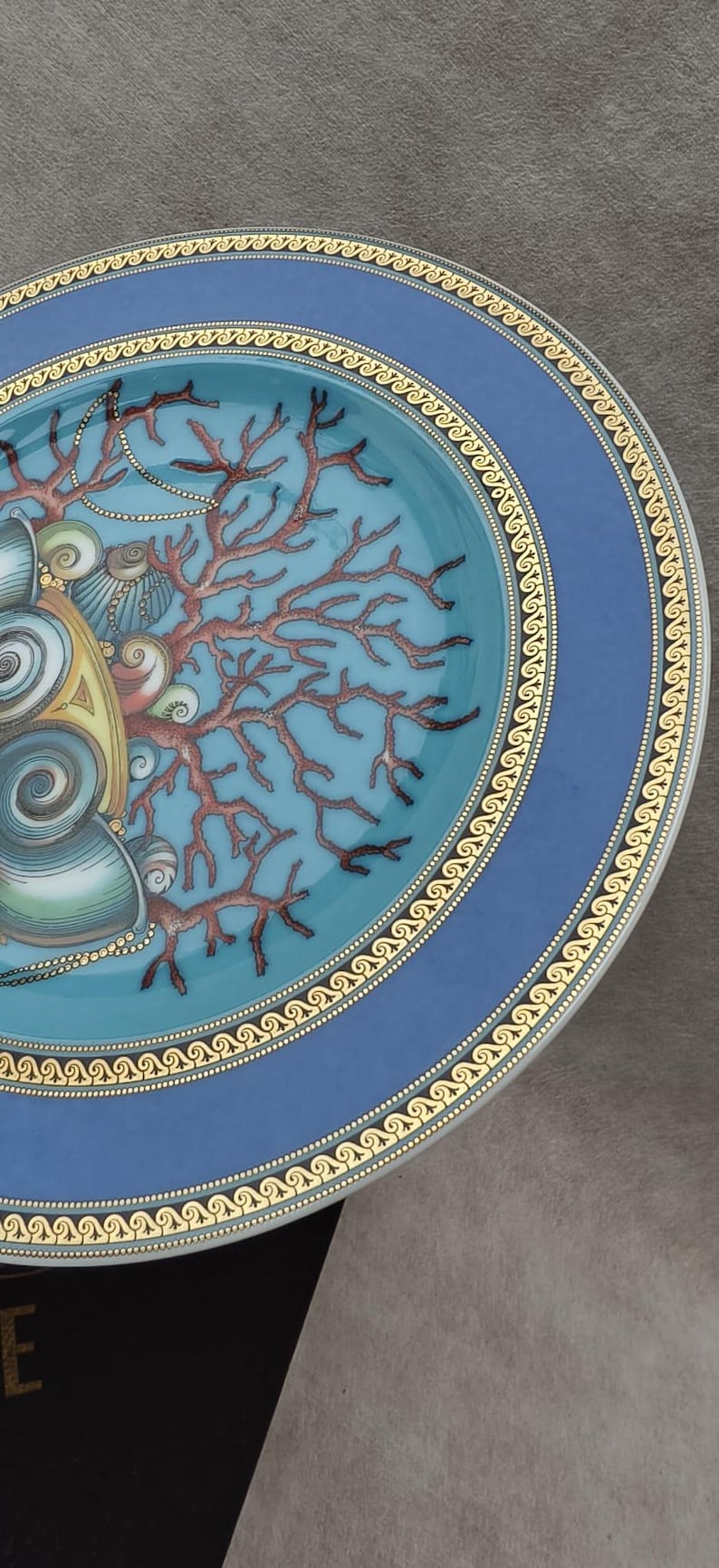 May include: Decorative ceramic plate with a blue and gold rim. The plate features a turquoise center with a coral and seashell design. The coral is brown, and the seashells are beige and gold.