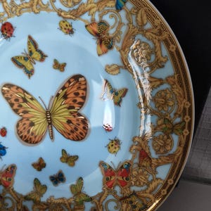 May include: A decorative plate with a light blue centre featuring colourful butterflies and ladybirds. The plate has a gold ornate border with a repeating pattern. The butterflies are in shades of orange, yellow, blue, and green.