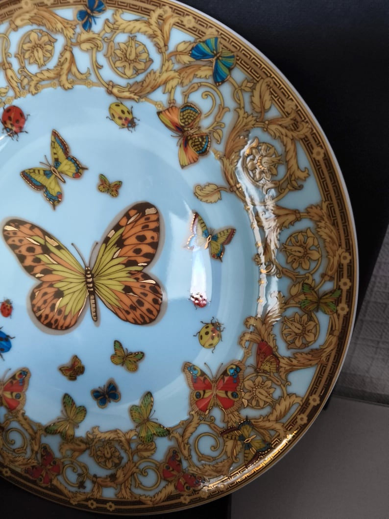 May include: A decorative plate with a light blue centre featuring colourful butterflies and ladybirds. The plate has a gold ornate border with a repeating pattern. The butterflies are in shades of orange, yellow, blue, and green.