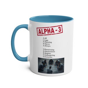 Op de afbeelding: Witte keramische mok met een blauwe binnenkant en handvat. De mok heeft de tekst "ALPHA-3" in een rood stempelachtig ontwerp, samen met een lijst met woorden en een foto van plannende mensen.