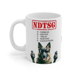 May include: White ceramic mug featuring a German Shepherd wearing sunglasses and holding gadgets. The mug has the text "NDTSG" in a red stamp design, with the acronym's meaning below. A fun and unique gift for dog lovers.
