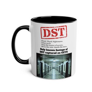 Puede incluir: Taza de cerámica blanca con asa e interior negros. La taza presenta el texto de sello rojo "DST" y una descripción de DST. Debajo del texto hay una foto de un largo pasillo.