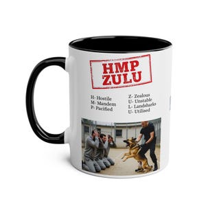 Op de afbeelding: Witte keramische mok met een zwart handvat en rand. De mok heeft de tekst "HMP ZULU" in een rood stempelachtig ontwerp. Daaronder staat een lijst met woorden en een foto van een hond en mensen.