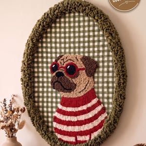 May include: An oval wall hanging featuring a punch needle pug wearing red-rimmed sunglasses and a red and white striped sweater. The pug is centered on a green and white gingham fabric background, framed by a thick, textured olive green yarn border.