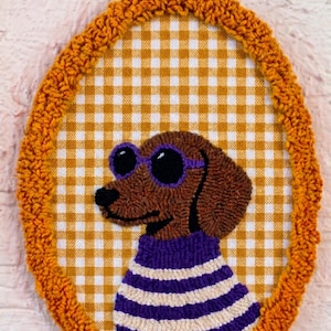 May include: An oval punch needle wall hanging featuring a brown dachshund wearing purple sunglasses and a purple and white striped sweater. The dog is centered on a yellow and white gingham fabric background, framed by a textured orange yarn border.