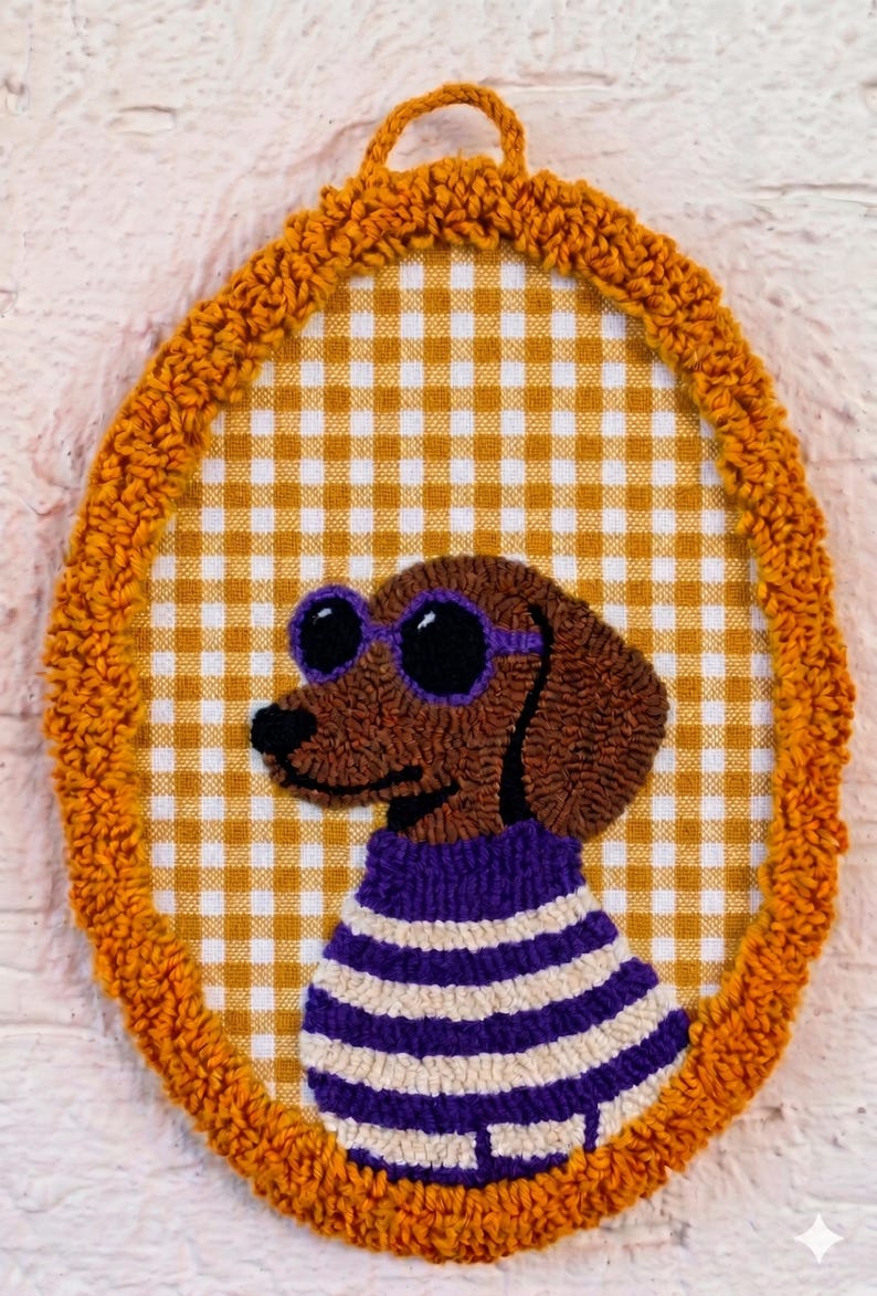 May include: An oval punch needle wall hanging featuring a brown dachshund wearing purple sunglasses and a purple and white striped sweater. The dog is centered on a yellow and white gingham fabric background, framed by a textured orange yarn border.