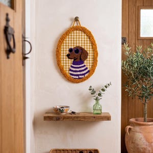 May include: An oval-shaped wall hanging featuring a tufted brown dachshund wearing purple sunglasses and a purple and white striped sweater. The background is a yellow and white gingham pattern, framed by a textured mustard yellow border.