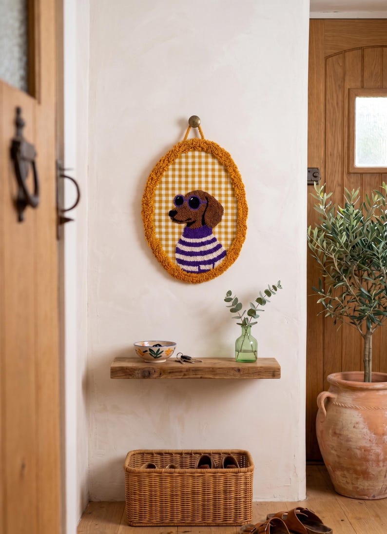 May include: An oval-shaped wall hanging featuring a tufted brown dachshund wearing purple sunglasses and a purple and white striped sweater. The background is a yellow and white gingham pattern, framed by a textured mustard yellow border.