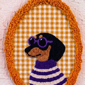 May include: An oval punch needle wall hanging featuring a black and tan dachshund wearing purple round glasses and a purple and white striped sweater. The dog is centered on a yellow and white gingham fabric background with a textured orange border.