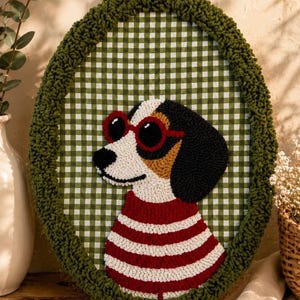 May include: An oval punch needle wall hanging featuring a dog wearing red-rimmed sunglasses and a red and white striped shirt. The dog is set against a green and white gingham background, framed by a thick, textured dark green yarn border.
