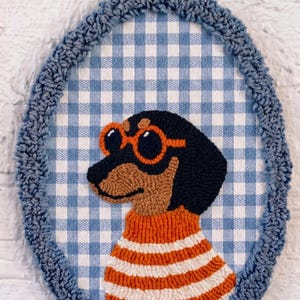 May include: An oval-shaped punch needle wall hanging featuring a black and tan dachshund wearing orange-rimmed sunglasses and an orange and white striped sweater. The dog is set against a blue and white gingham background with a blue yarn border.
