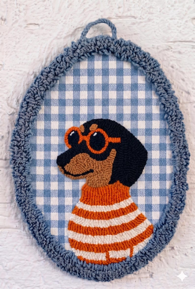 May include: An oval-shaped punch needle wall hanging featuring a black and tan dachshund wearing orange-rimmed sunglasses and an orange and white striped sweater. The dog is set against a blue and white gingham background with a blue yarn border.