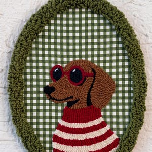 May include: An oval-shaped punch needle wall hanging featuring a brown dachshund wearing red-rimmed sunglasses and a red and white striped sweater. The dog is set against a green and white gingham fabric background, framed by a thick olive green border.