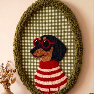 May include: An oval wall hanging featuring a punch needle dachshund wearing red-rimmed sunglasses and a red and white striped sweater. The dog is set against a green and white gingham fabric background, framed by a thick, textured olive green yarn border.
