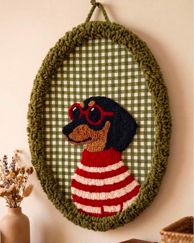 May include: An oval wall hanging featuring a punch needle dachshund wearing red-rimmed sunglasses and a red and white striped sweater. The dog is set against a green and white gingham fabric background, framed by a thick, textured olive green yarn border.