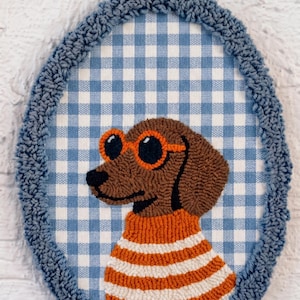 May include: An oval punch needle wall hanging featuring a brown dog wearing orange-rimmed sunglasses and an orange and white striped sweater. The dog is set against a blue and white gingham fabric background, framed by a textured blue yarn border.