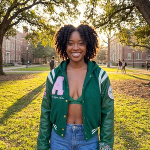 Op de afbeelding: Een groene varsity jas met witte strepen en een roze "A". De jas heeft groene leren mouwen, gedragen met een groene bh en een spijkerbroek. De achtergrond toont een universiteitscampus.