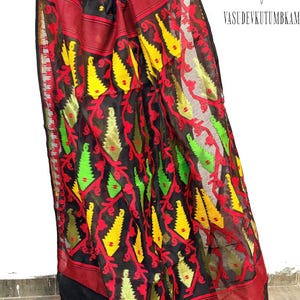 May include: A black sari with a vibrant pattern of red, yellow, and green floral and geometric designs. The sari has a red border and is made of a sheer fabric. The text "VASUDEVKUTUMBKAM" is visible in the image.