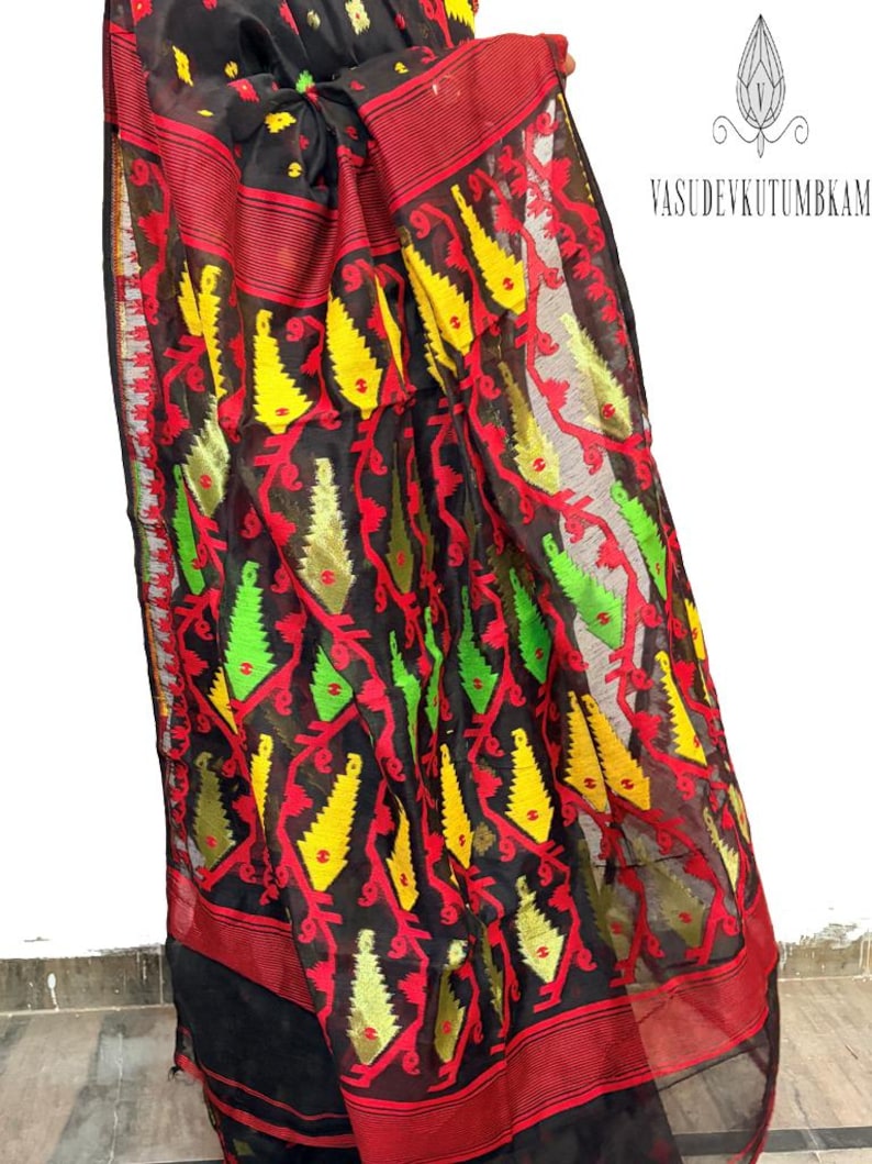 May include: A black sari with a vibrant pattern of red, yellow, and green floral and geometric designs. The sari has a red border and is made of a sheer fabric. The text "VASUDEVKUTUMBKAM" is visible in the image.