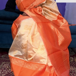 May include: A cream-colored sari with an orange border and accents. The sari features a gold peacock pattern and decorative gold trim. The text "VASUDEVKUTUMBKAM" is visible in the upper right corner.