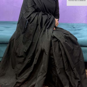 May include: A black saree with a matching black top. The saree is draped over a person sitting on a teal couch. The saree has a textured appearance and a fringe with tassels. The background is a purple wall.