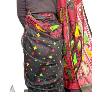 May include: A black sari with a vibrant red, yellow, and green patterned border and accents. The sari features a repeating pattern of colorful floral and geometric shapes. The fabric appears sheer and lightweight, with a decorative edge.