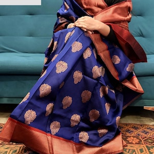May include: A blue saree with gold floral patterns and a red border. The saree is draped over a teal couch. The saree has a rich, textured appearance, with intricate detailing in the gold patterns.